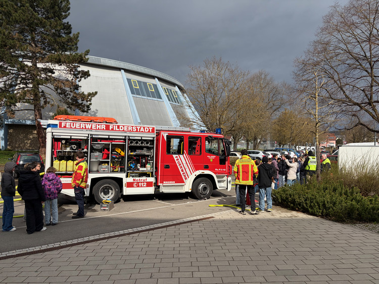 Feuerwehrauto steht geöffnet da, Feuerwehrmänner und Schüler stehen daneben und machen Löschübungen