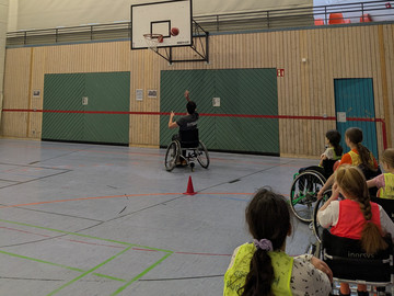 Mann im Rollstuhl wirft einen Basketball Richtung Korb, am Rand sitzen Schüler in Rollstühlen und beobachten ihn