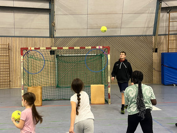 Turnhalle, drei jüngere Schülerinnen schießen auf Tor, ein älterer Schüler steht am Tor