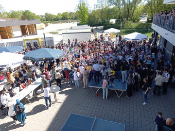 große Gruppe von Menschen auf dem Schulhof bei sonnigem Wetter, Sonnenschirme, Tische